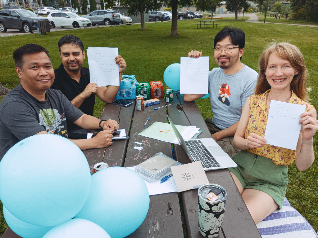 Photo of some participants around a picnic table decorated with green-ish balloons, and people hold handwritten pages of HTML to the camera.